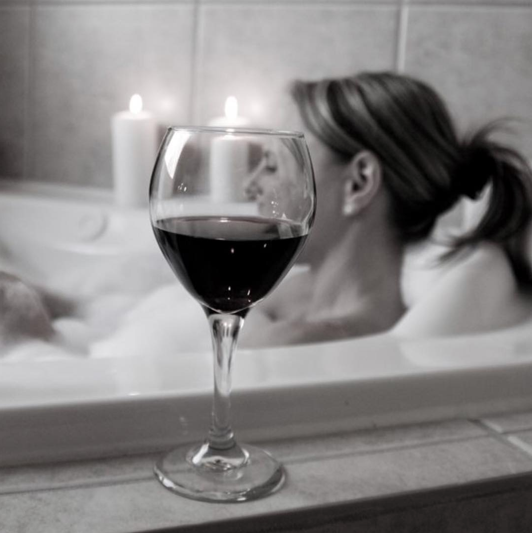 Black and white photo of a glass of red wine on a bathtub edge with candles