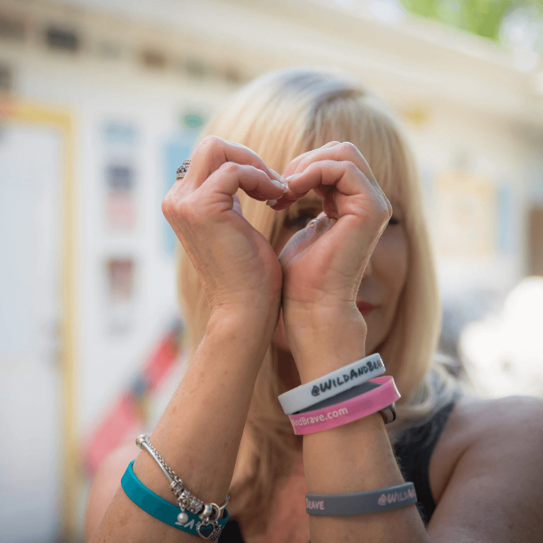 A blonde woman forming a heart shape with her hands, wearing Wild + Brave wristbands