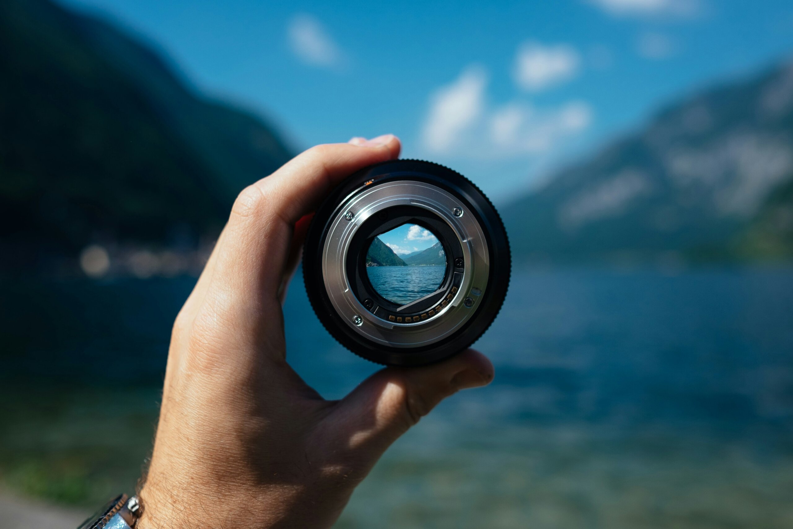 Hand holding a camera lens up to a mountain lake