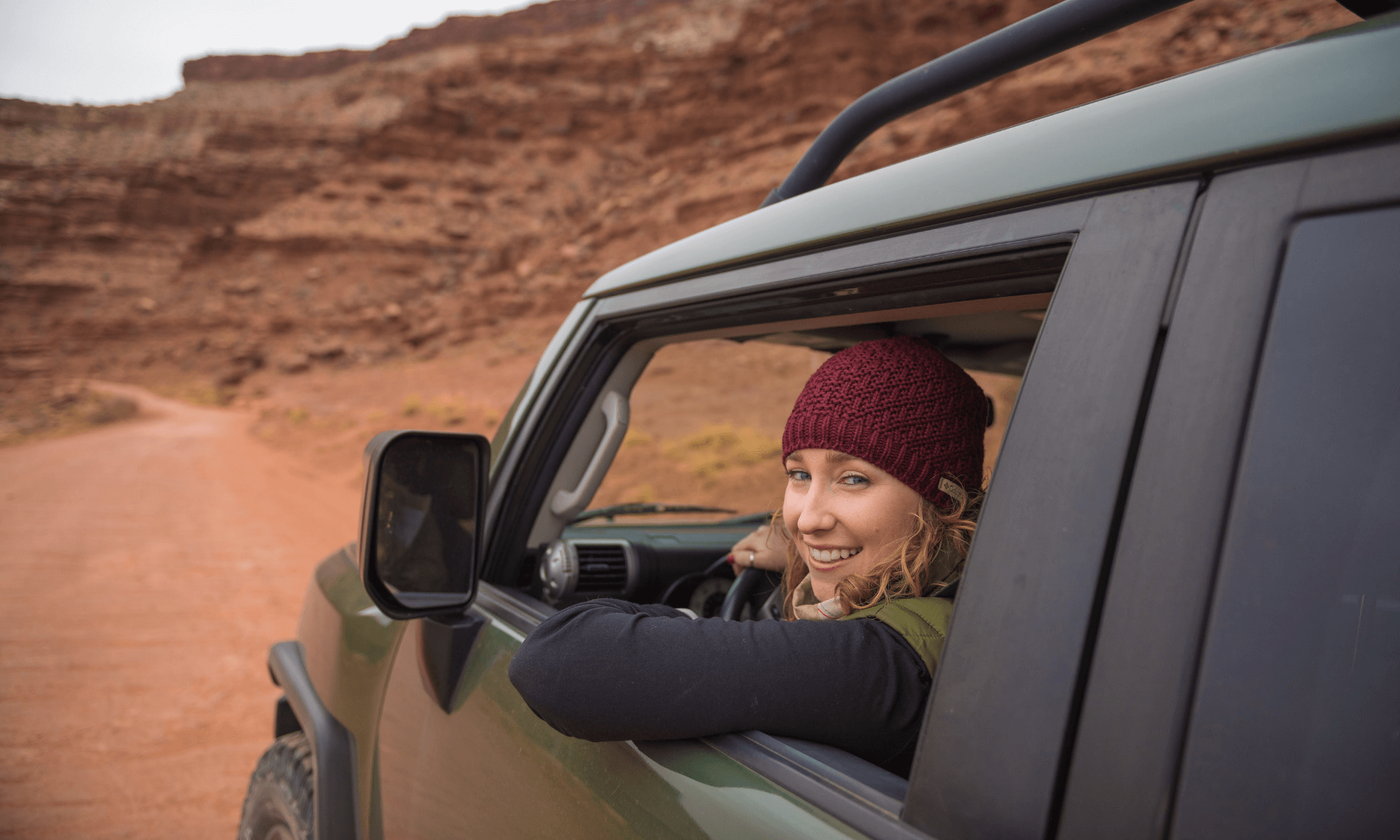 Smiling woman in a red beanie leaning out of a green SUV window on a red-dirt desert road