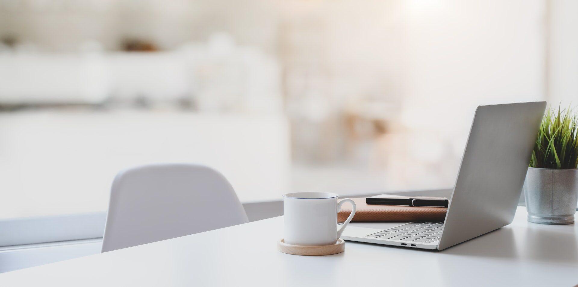 Bright minimalist white desk with an open laptop, coffee mug, notebook and small potted plant
