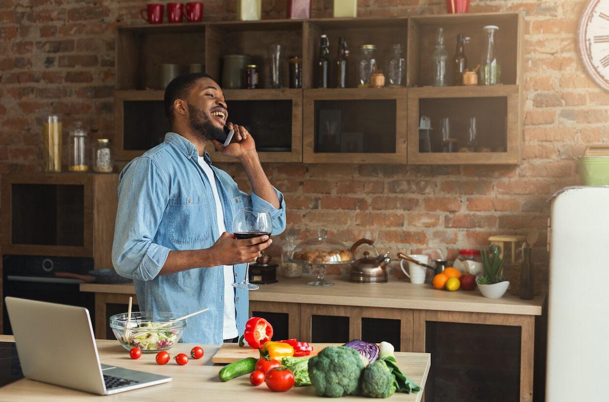 A Black man laughing on the phone with a wine glass in hand, standing in a rustic kitchen with fresh vegetables on the counter