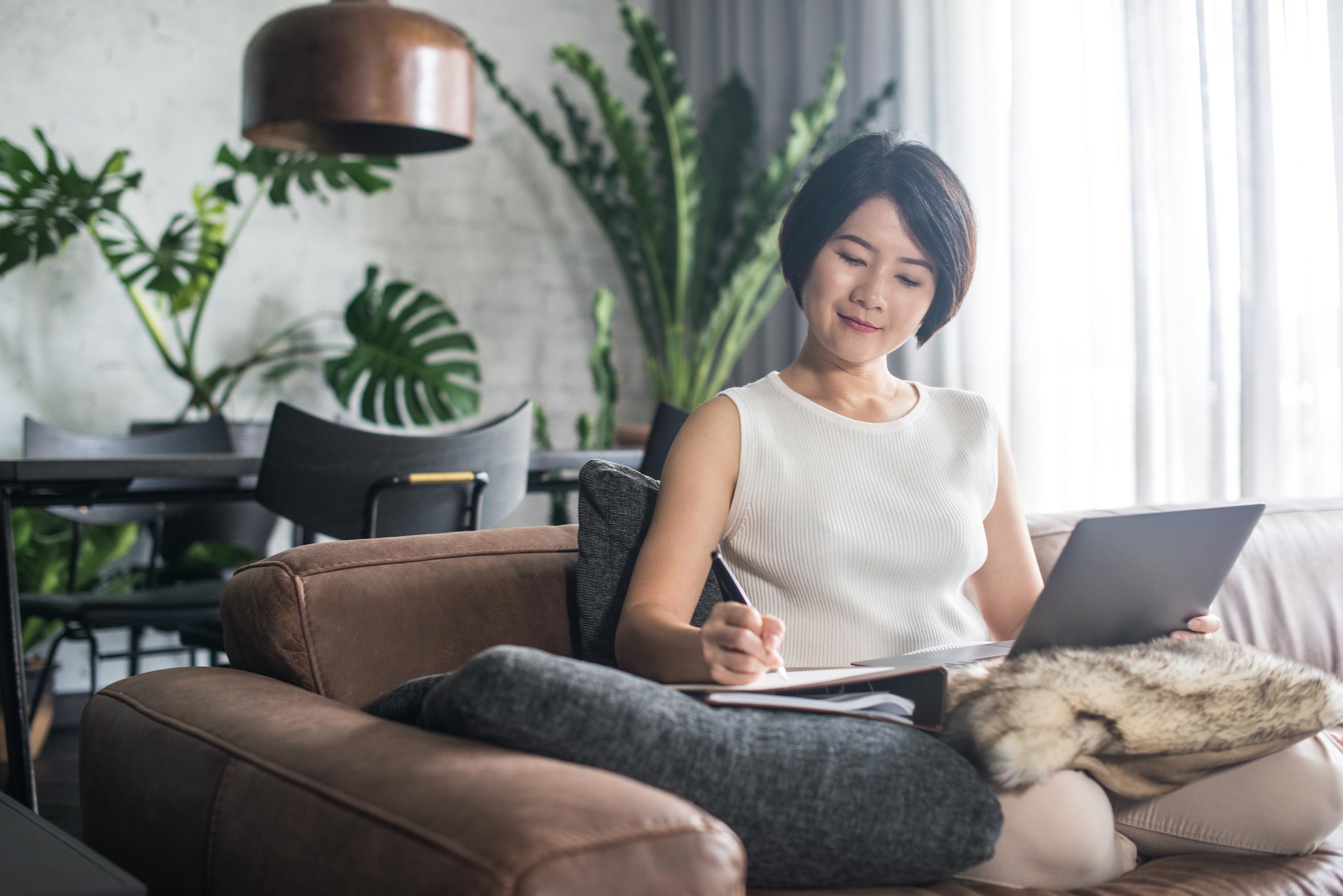 A young Asian woman sitting on a sofa journaling in a notebook while holding a tablet, plants behind her
