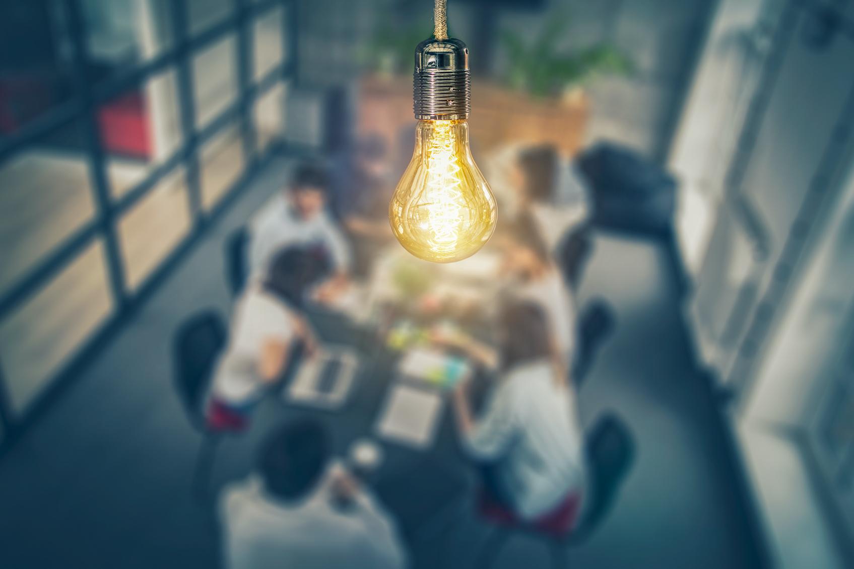 A glowing Edison lightbulb hanging above a blurred group of people working together at a table
