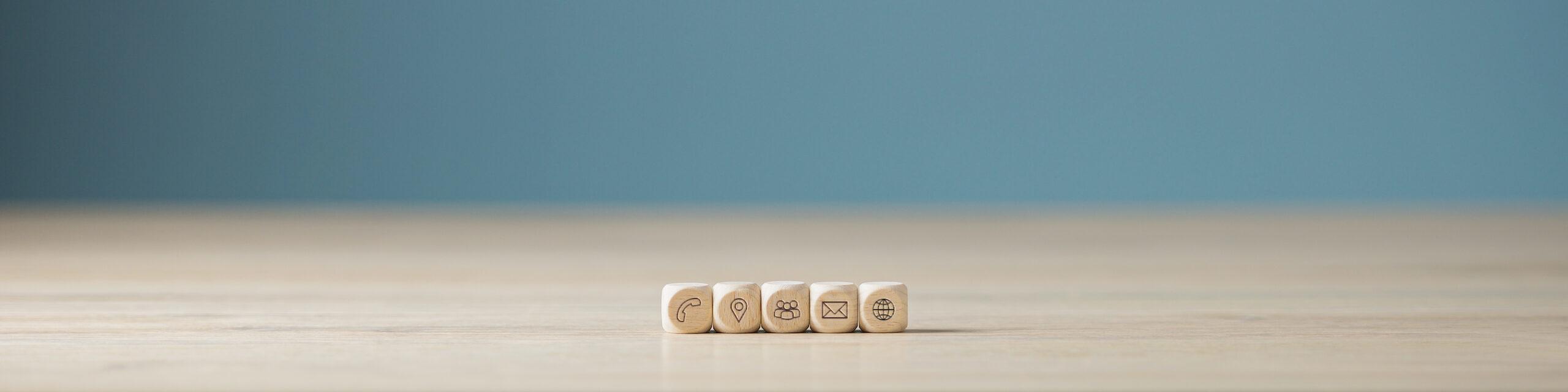Five wooden blocks engraved with phone, location pin, people, email and globe icons on a light wood surface against a teal background