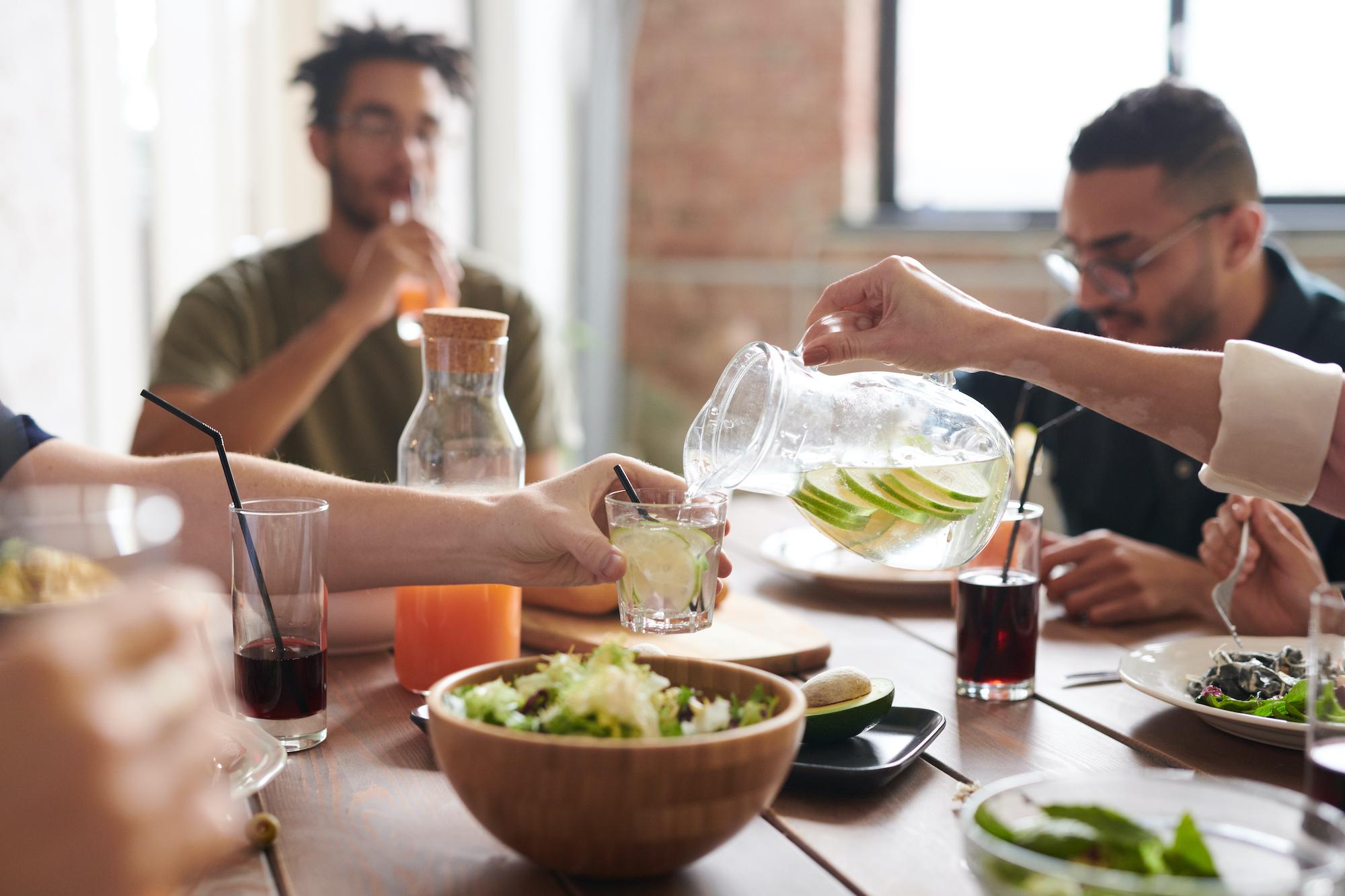A diverse group of people sharing a healthy meal at a wooden table while someone pours infused water