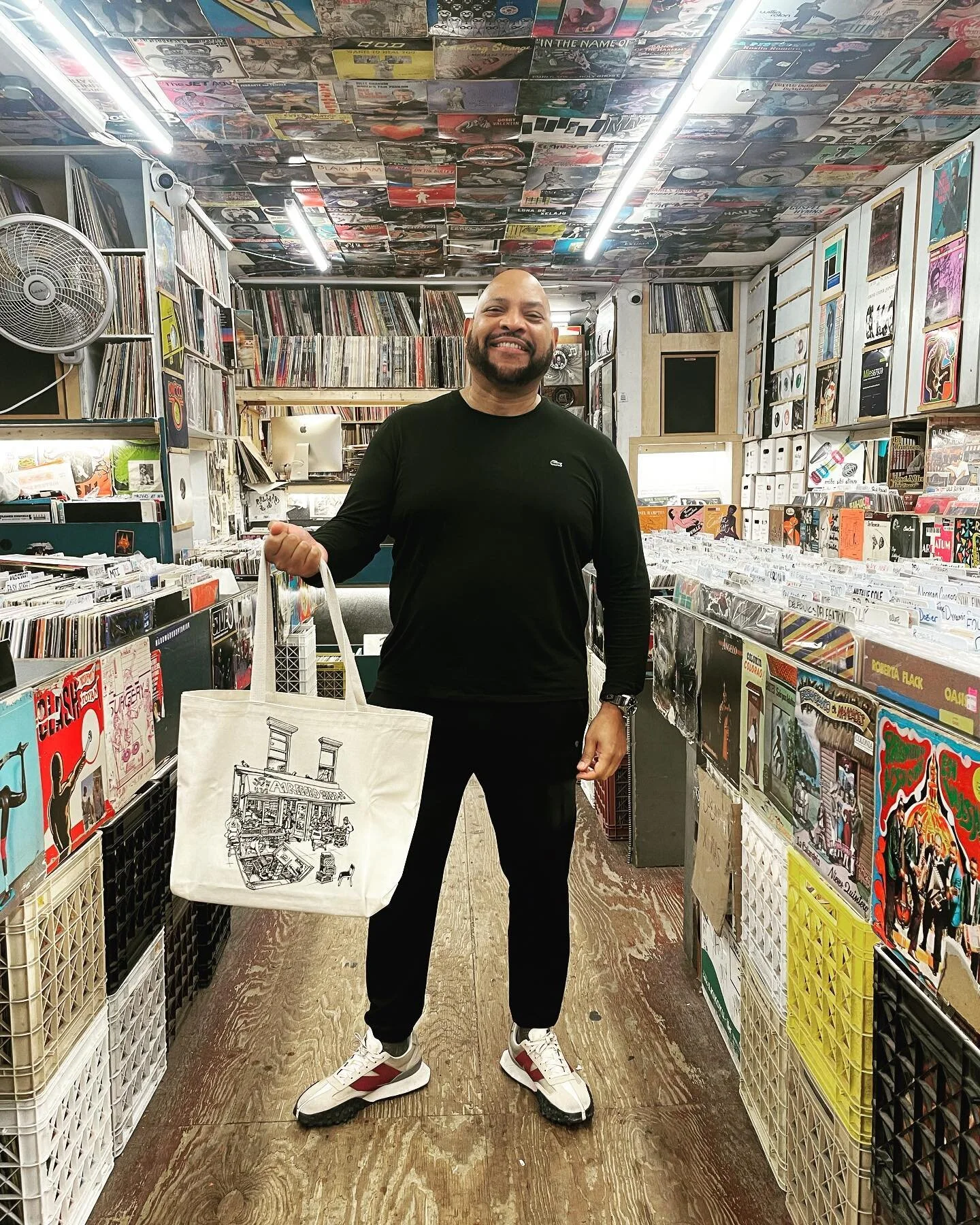 Man in a black Lacoste outfit standing in the record shop aisle holding an A-1 Record Shop tote bag