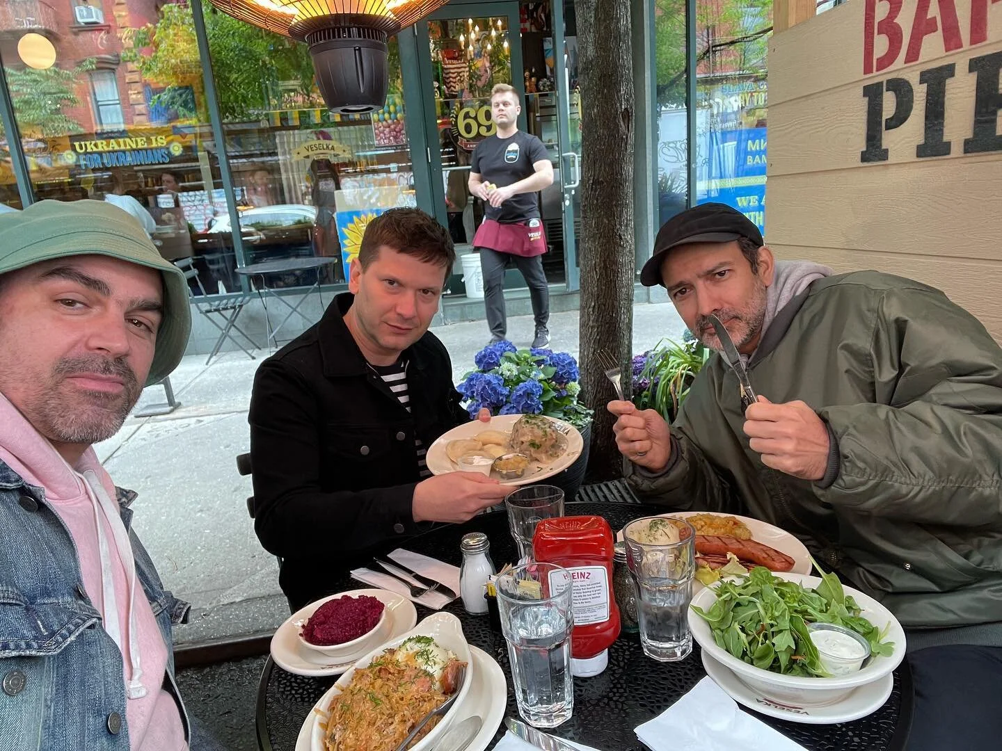 Three men at an outdoor café table near the shop with Veselka in the background