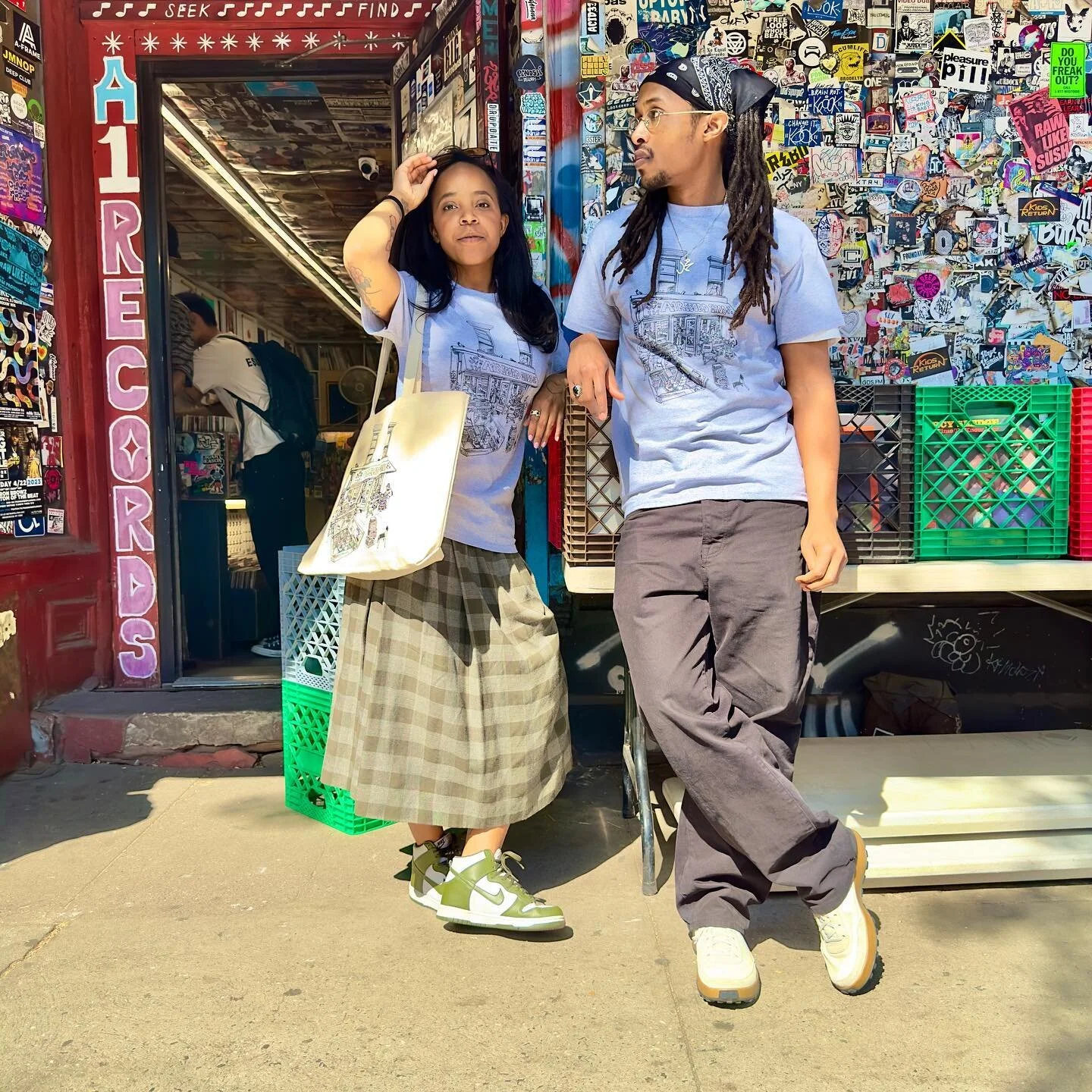Two people in matching A-1 Record Shop t-shirts posing outside the store's sticker-covered wall