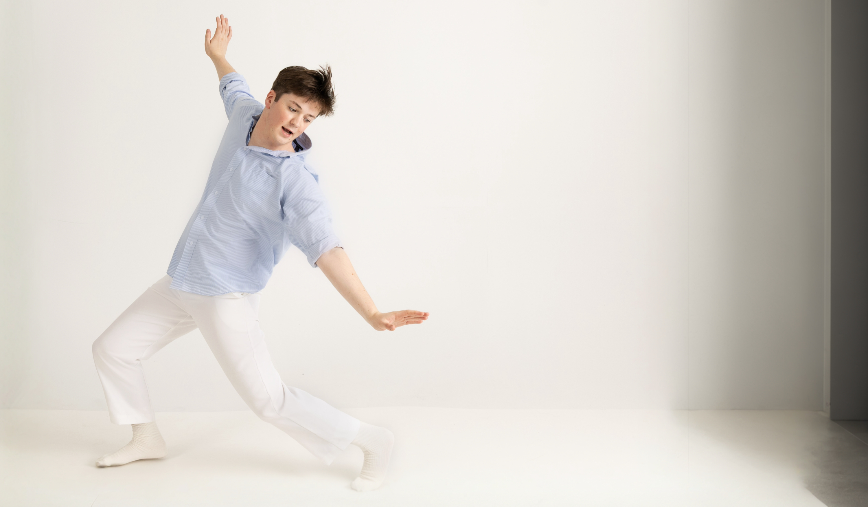 Benji Godley-Fisher performing a dynamic dance pose in a light blue shirt and white pants against a white studio background