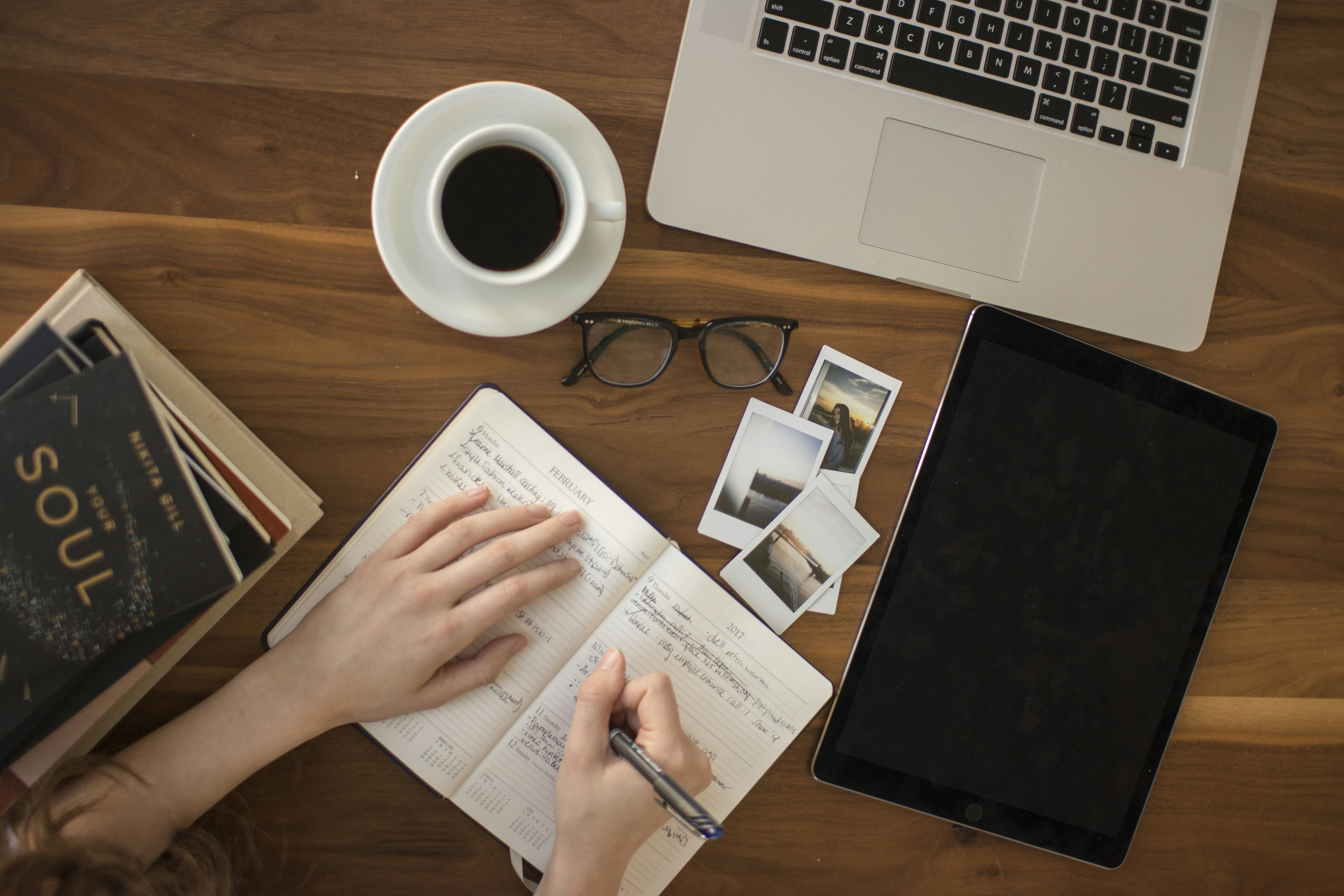 Overhead flat-lay of a wooden desk with black coffee, an open journal, laptop, tablet, glasses and Polaroid photos