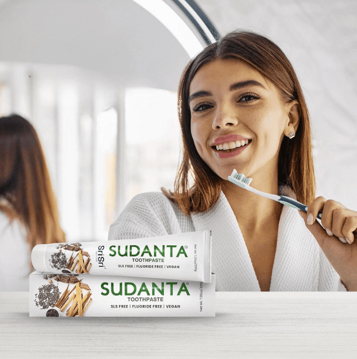 A smiling woman holding a toothbrush beside Sudanta Toothpaste boxes — a wellness oral-care brand now stocked in TJX.