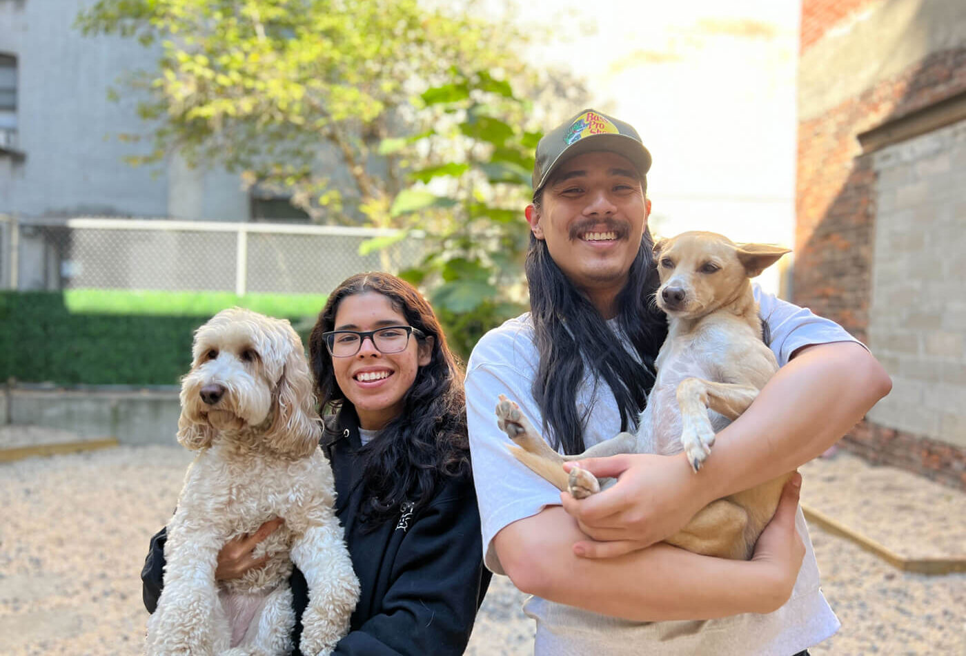 Two smiling DOGMA staff members holding a white labradoodle and a tan Chihuahua in the outdoor yard