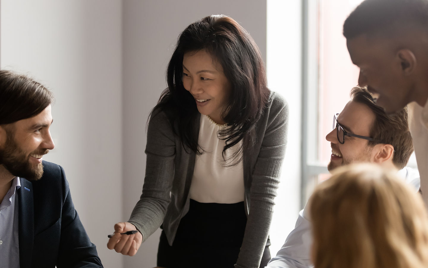 Diverse group of four colleagues laughing and collaborating around a table, a woman in a gray blazer leading the discussion