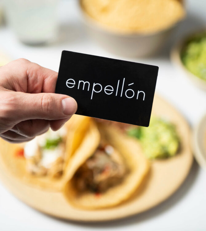 A hand holds a black Empellón gift card above a plate of tacos.