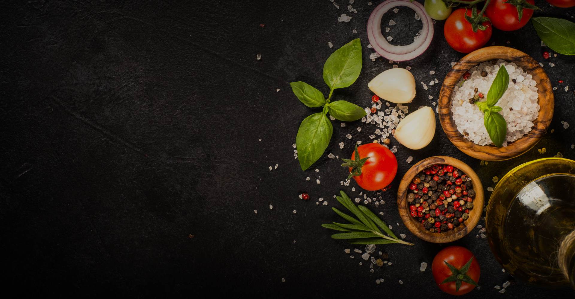 Flat-lay of basil, cherry tomatoes, garlic, salt in wooden bowls, peppercorns and olive oil on a dark surface