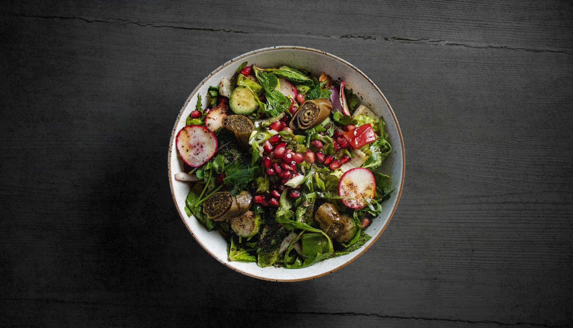 Overhead view of a Lebanese fattoush salad with pomegranate seeds, radishes, cucumber and rolled grape leaves on dark wood