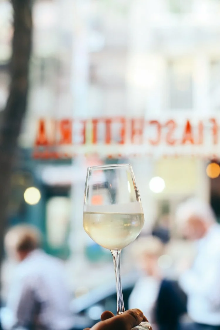 Hand holding a glass of white wine with the blurred Fiaschetteria sign behind