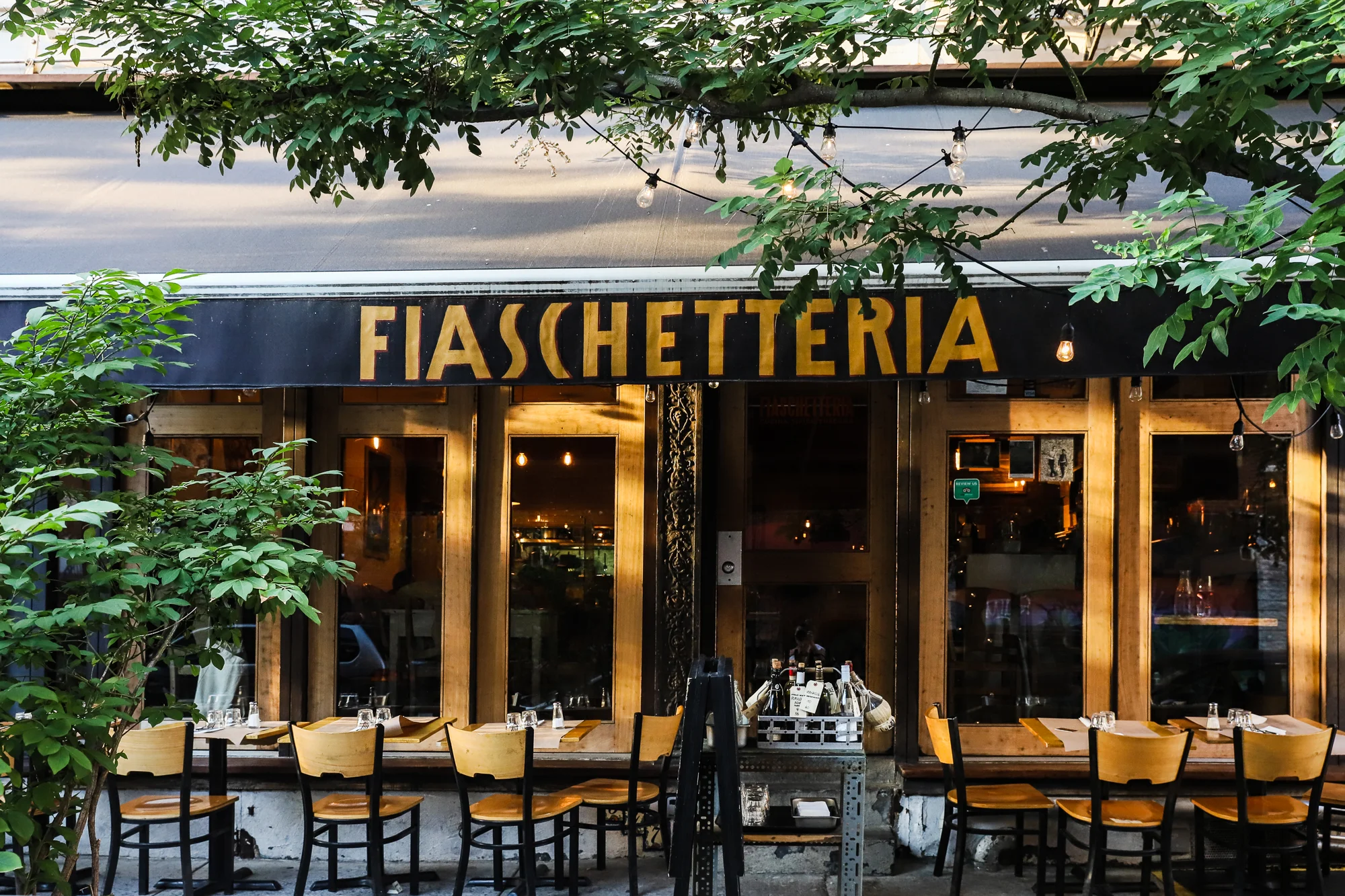 Facade with gold Fiaschetteria lettering on a black awning, sidewalk seating under trees and string lights