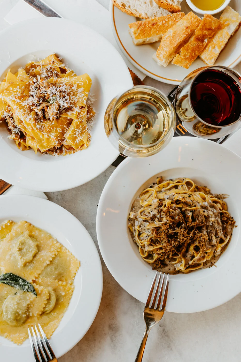 Overhead flat-lay of pappardelle, tagliatelle and ravioli with wine glasses on a marble table
