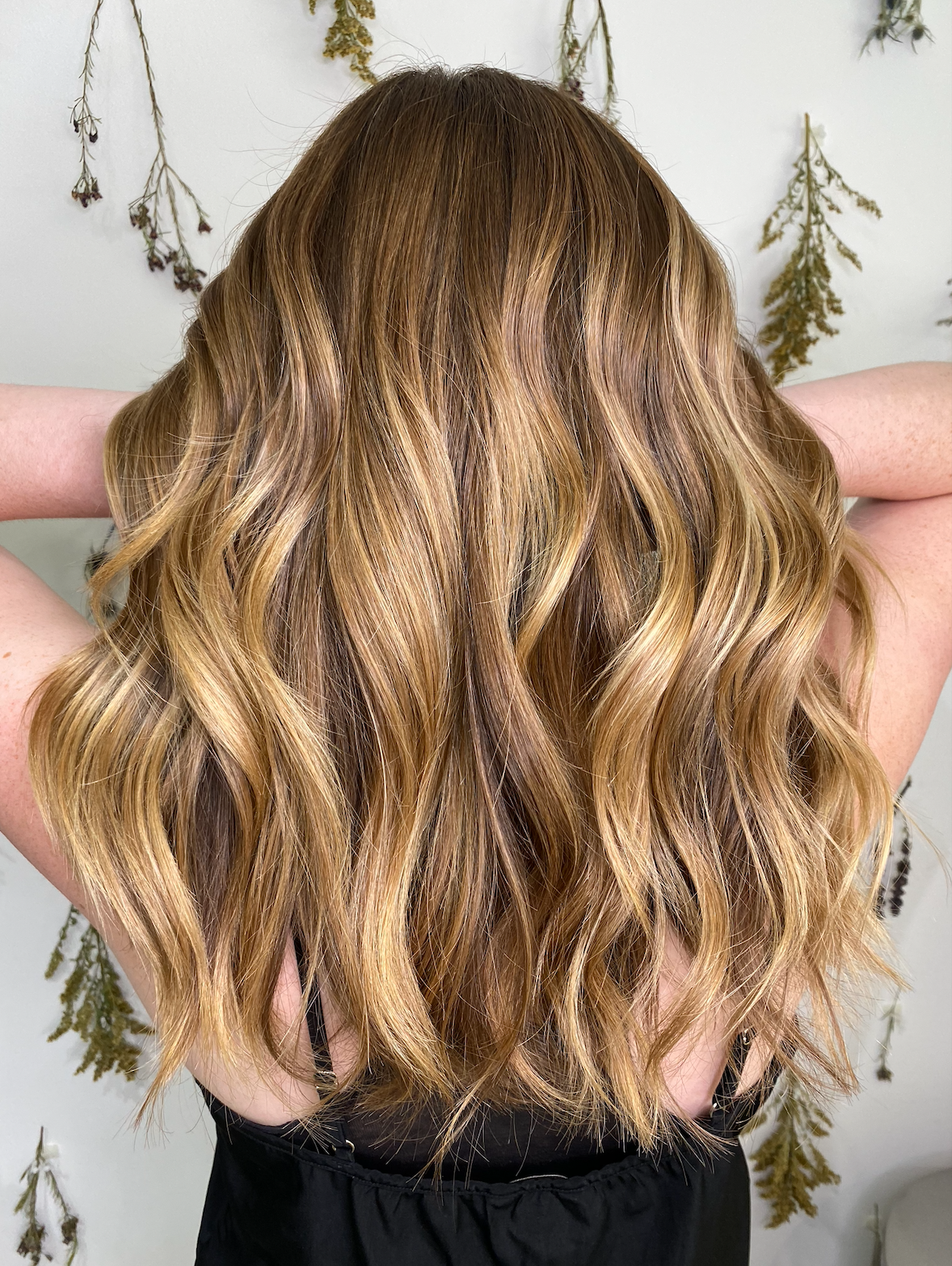 Long wavy golden-brunette balayage seen from behind against the dried flower wall at Green Velvet Parlor