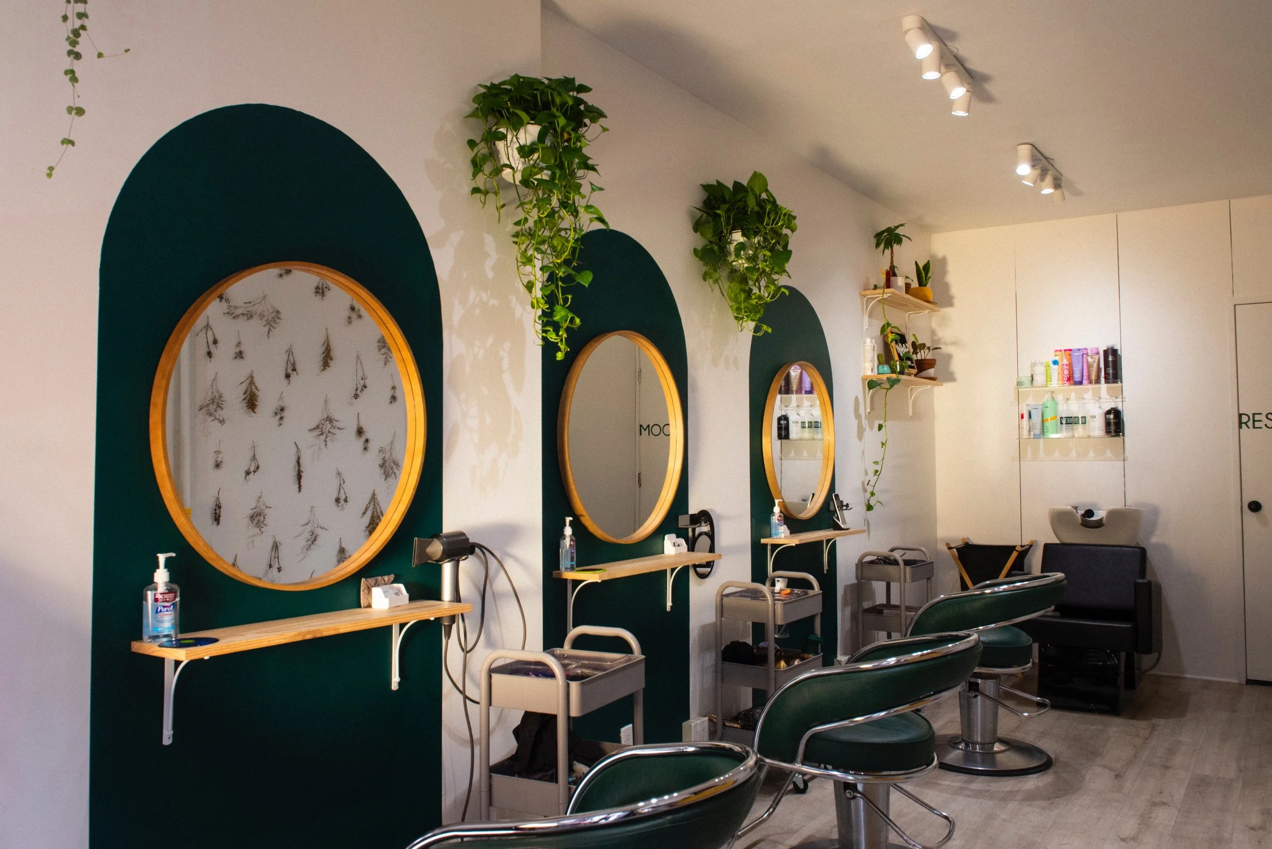 The Green Velvet Parlor salon interior: three styling stations against deep green arched wall panels with round gold-framed mirrors and hanging plants