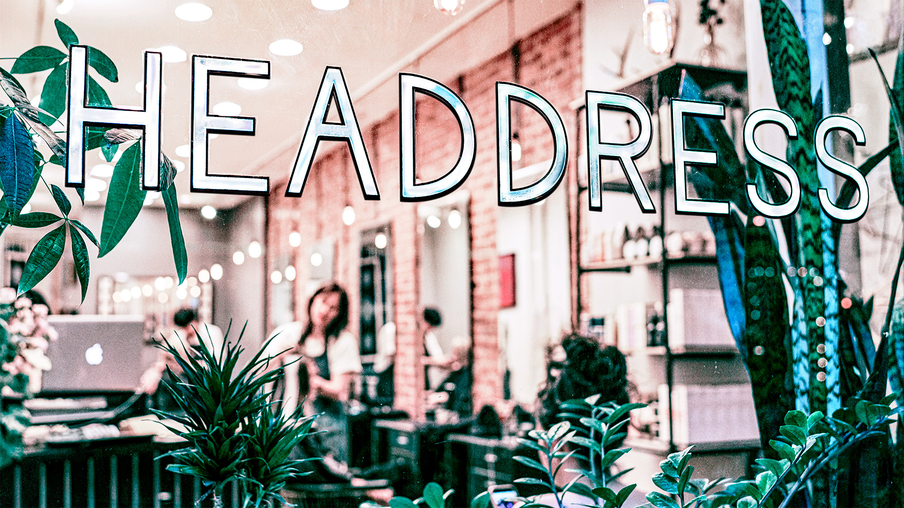 Headdress Hair Salon storefront window with illuminated HEADDRESS signage, plants in the foreground and the salon interior visible behind the glass