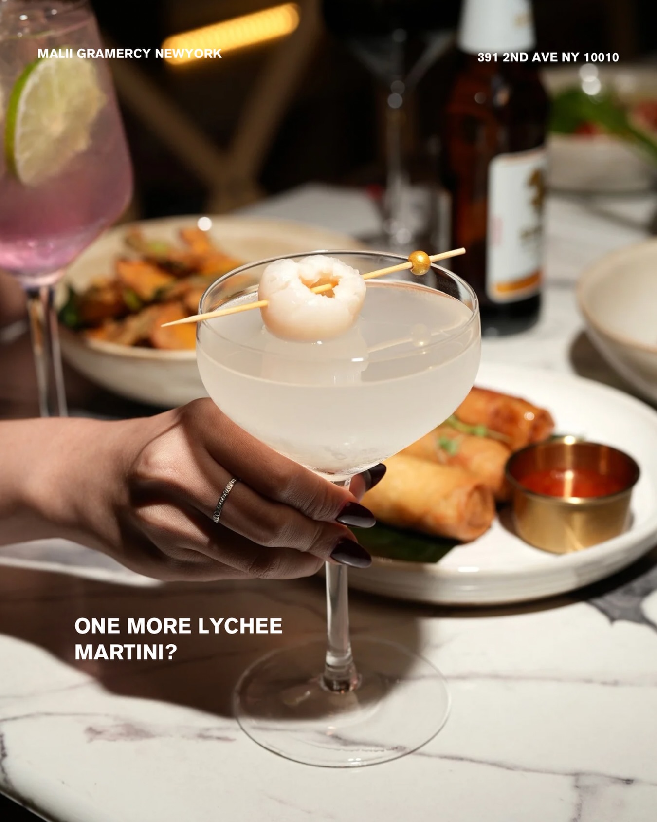 Hand holding a lychee martini garnished with a shrimp skewer, spring rolls behind