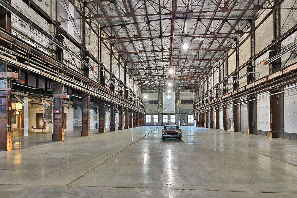 Massive industrial warehouse interior with steel truss structure, polished concrete floor and a classic car parked in the middle