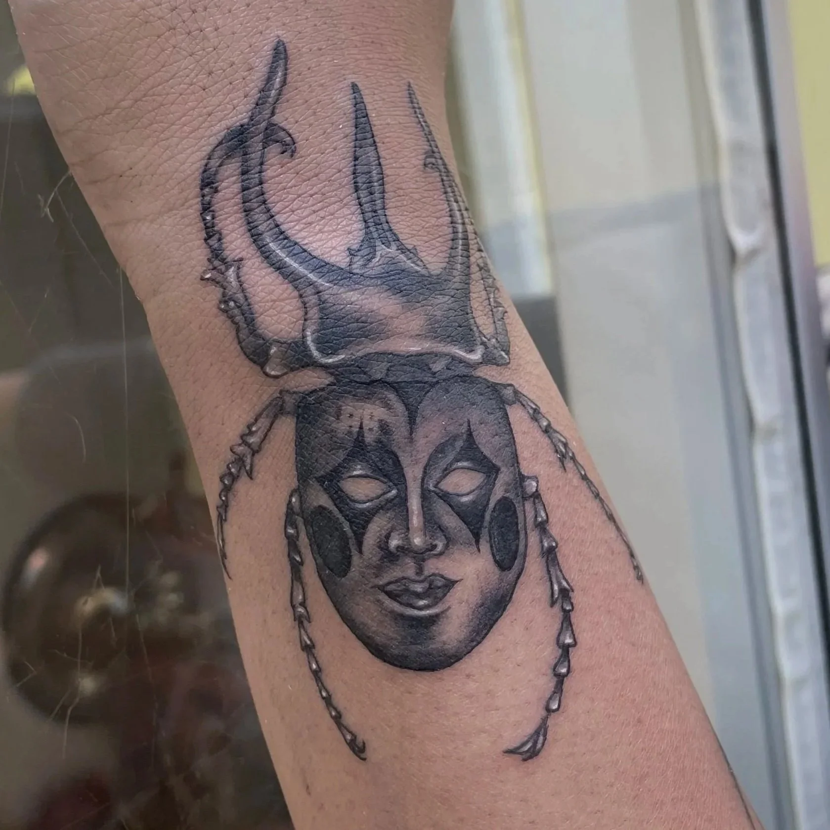Black-and-grey tattoo of a beetle with a theatrical mask for a face, on a wrist
