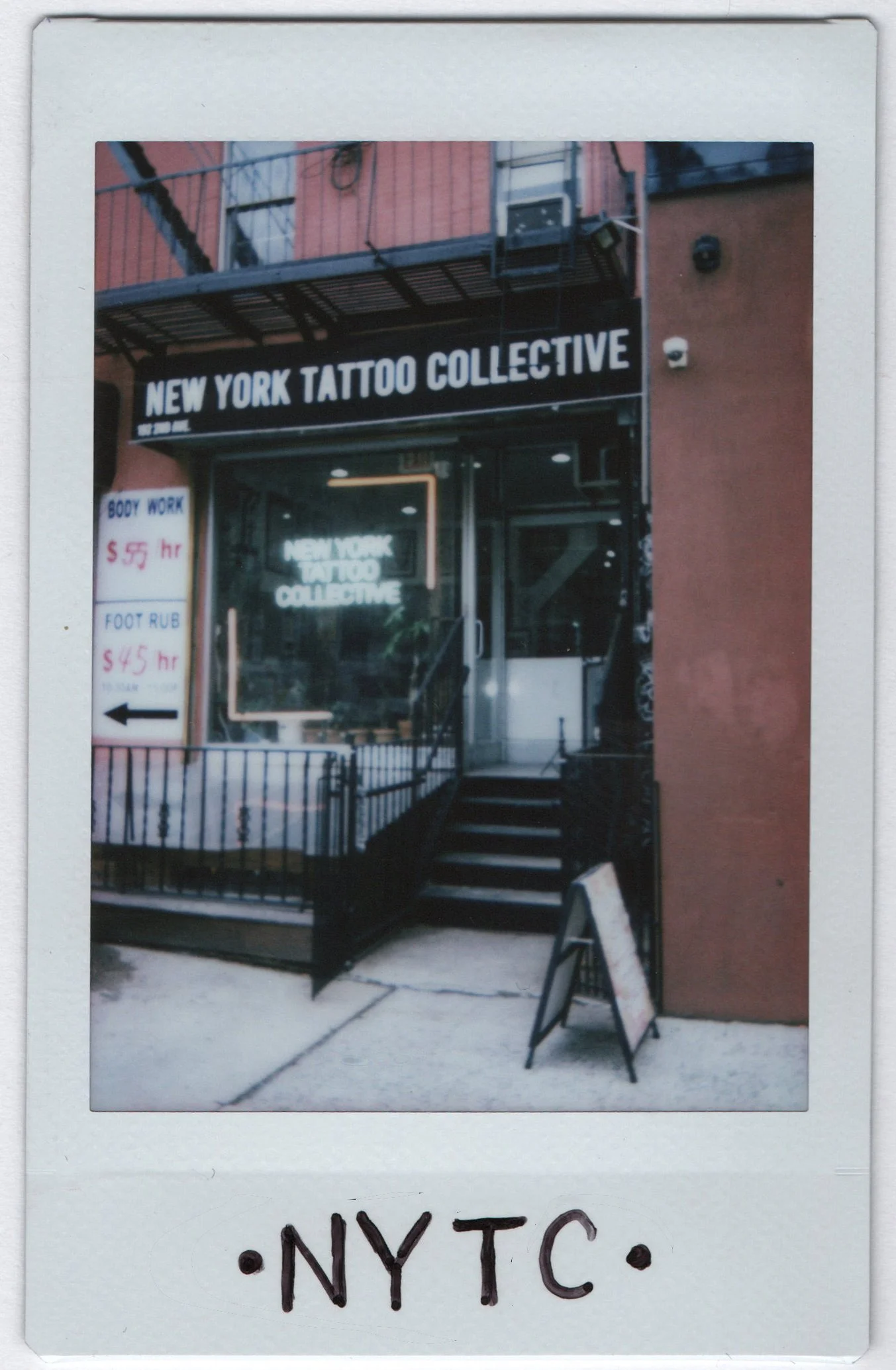 Polaroid-style photo of the New York Tattoo Collective storefront on 2nd Avenue, with the shop awning, neon window sign and stoop railing, and ·NYTC· written on the white border