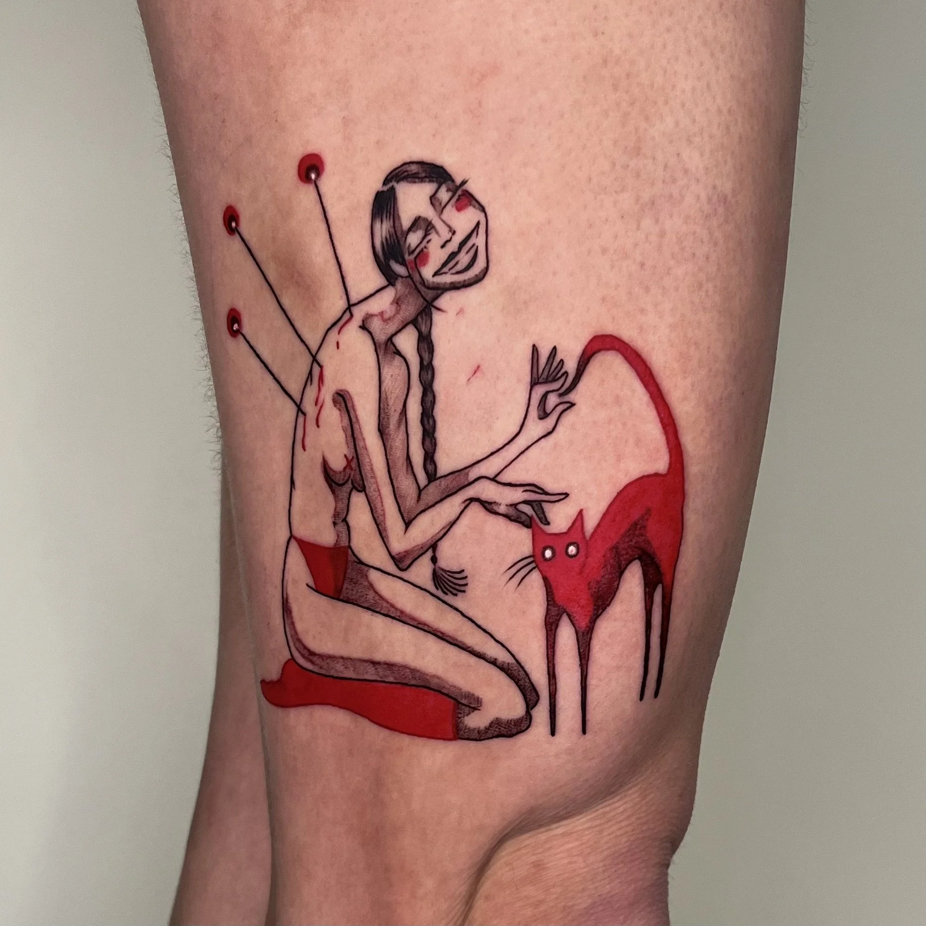 Illustrative black-and-red tattoo of a figure stuck with pins beside a red cat, on a leg