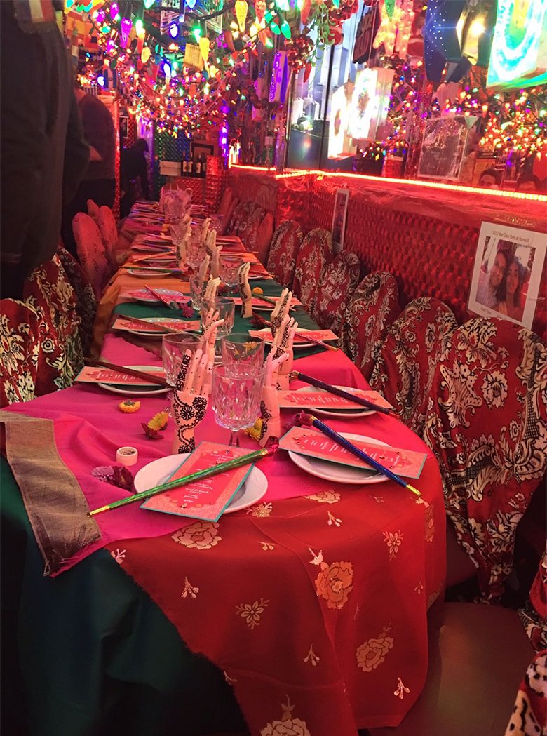 Long table set for a party at Panna II with red floral chair covers under a canopy of colored lights