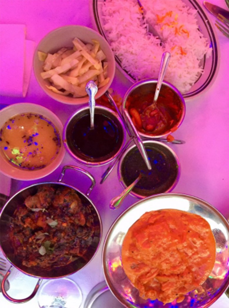 Overhead view of curries, rice and chutneys in steel bowls under pink light