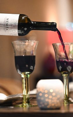 Red wine poured from a Nemea bottle into a goblet with candlelight behind
