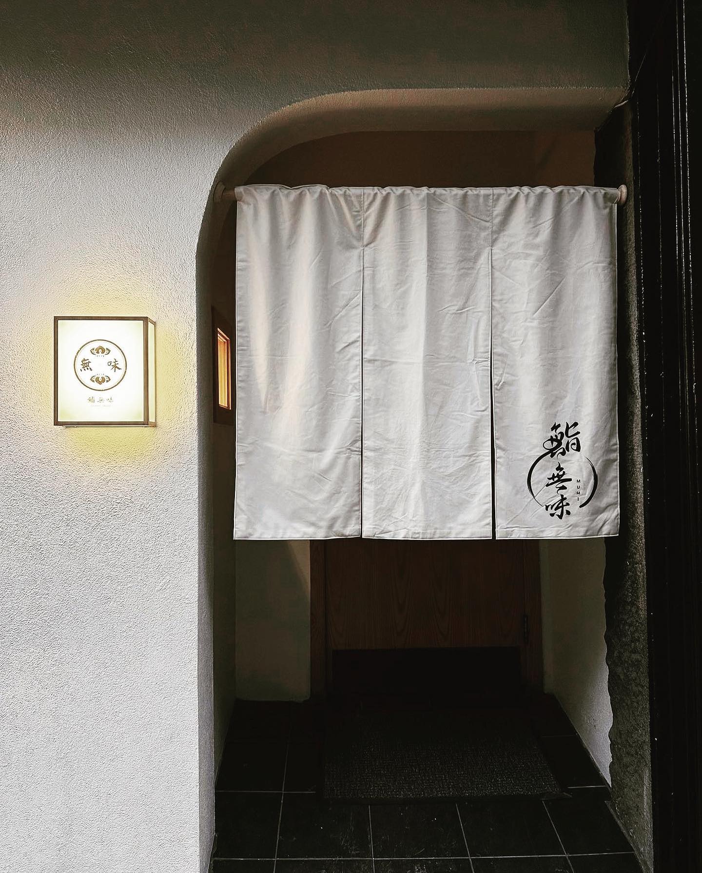 The entrance to Sushi Mumi — a white noren curtain hand-lettered with 鮨無味 hanging in an arched doorway, beside an illuminated lantern sign on a textured plaster wall.