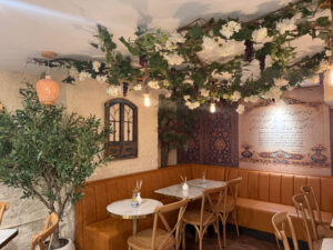 Corner booth beneath a floral ceiling installation, with a potted olive tree and arched window