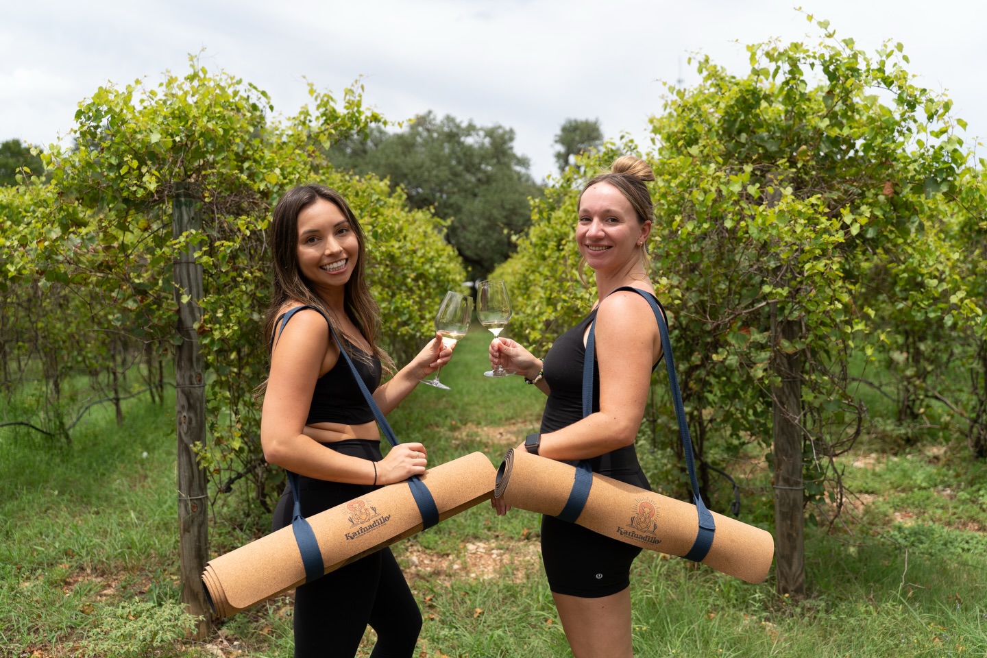 Two women toasting white wine in vineyard rows with cork yoga mats