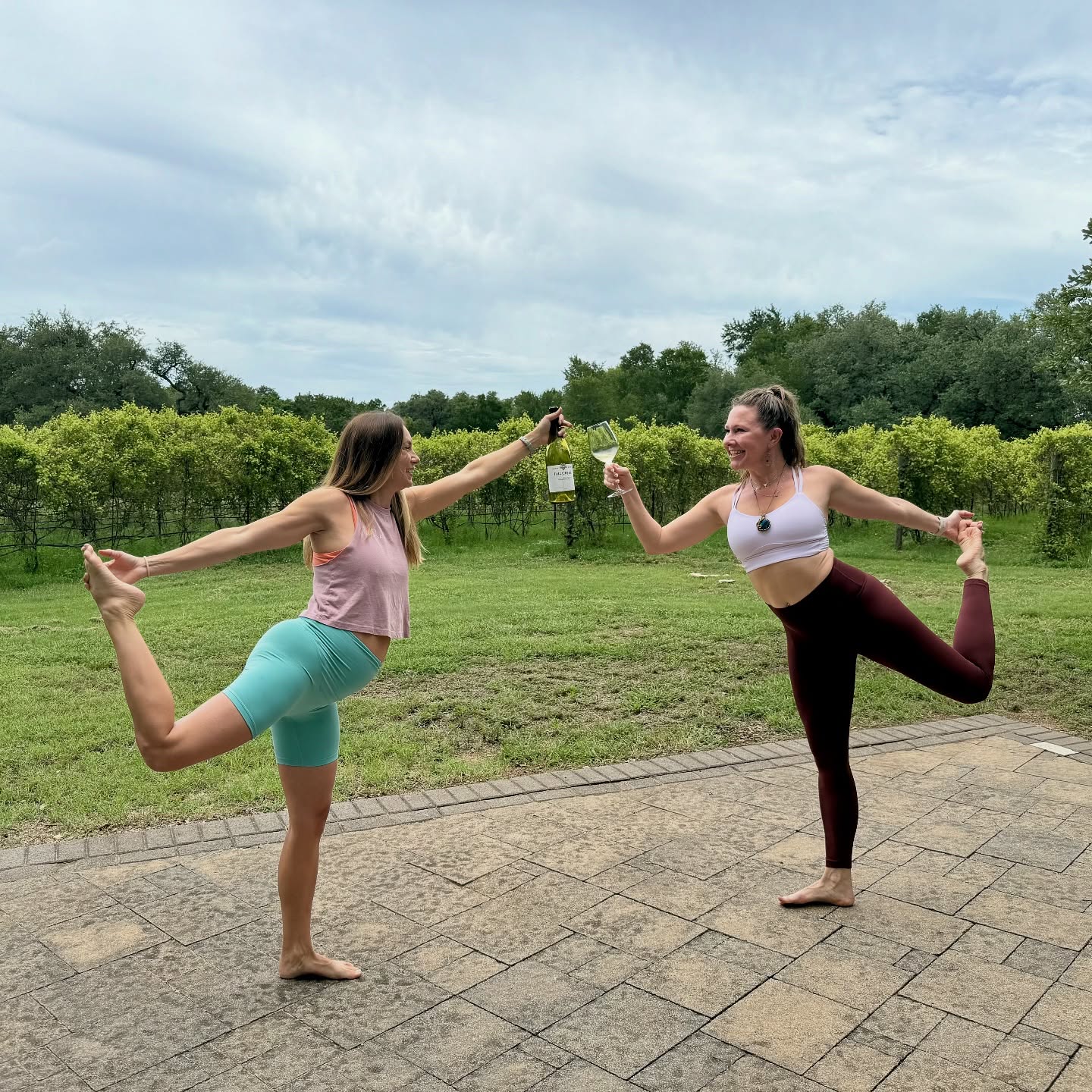 Two women balancing in a standing pose toasting wine at a vineyard