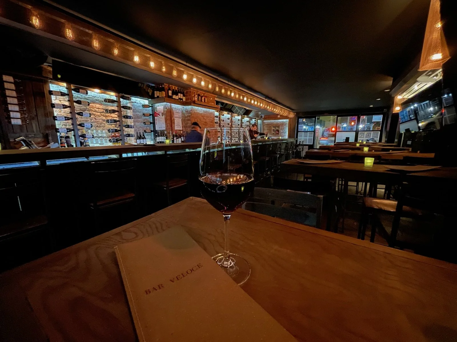 Bar Veloce SoHo interior — a Bar Veloce menu and a glass of red wine in the foreground, lit wine rack behind