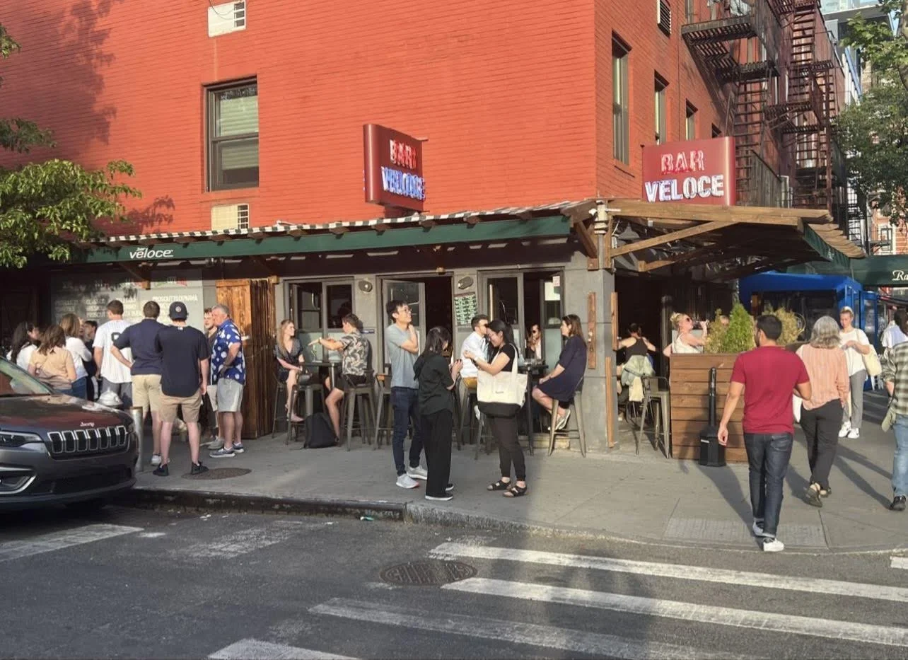 Bar Veloce exterior on a New York street — red brick facade, signage and a crowd of patrons at the sidewalk seating