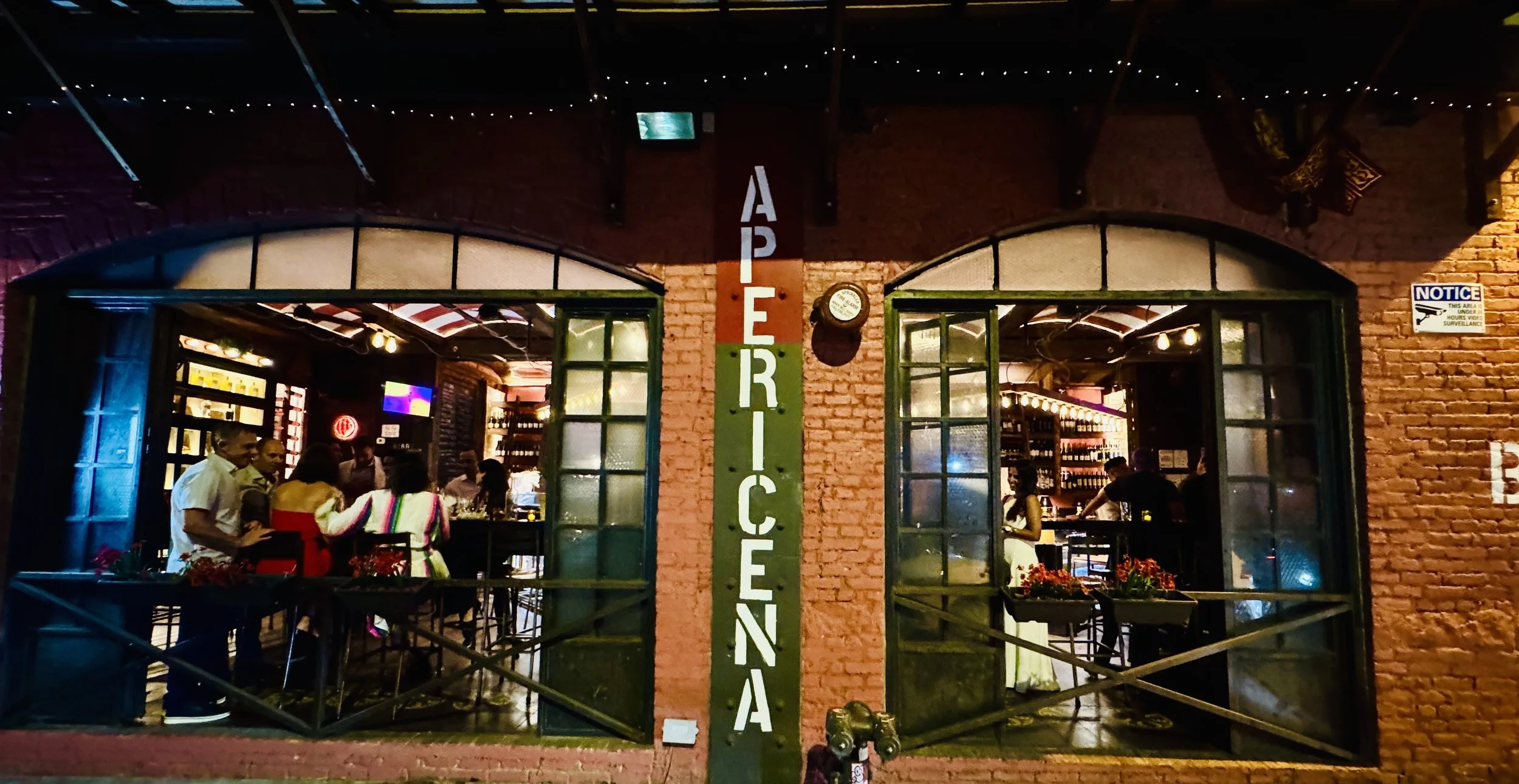 Apericena brick facade at night with two arched windows and an illuminated vertical sign
