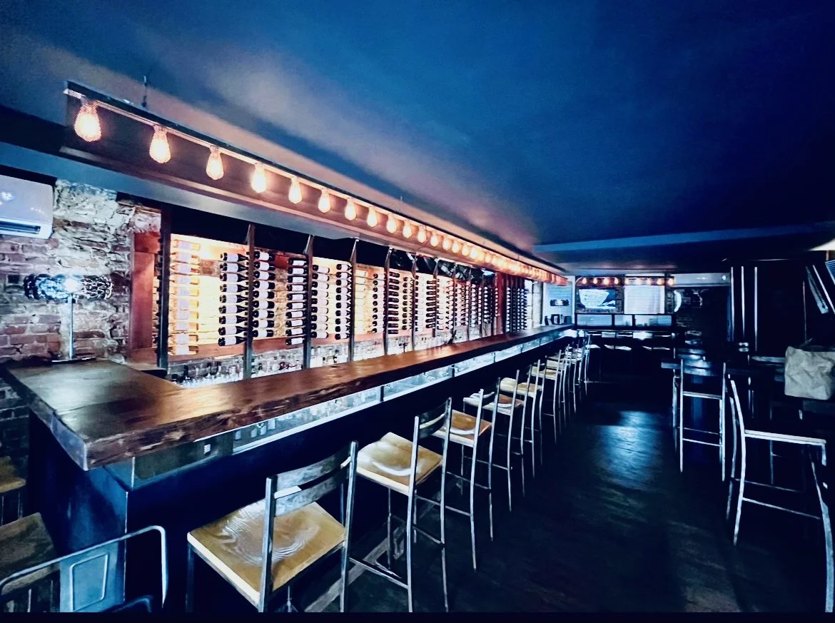Bar Veloce Chelsea — long dark bar with an illuminated wine rack wall and a strip of Edison pendants