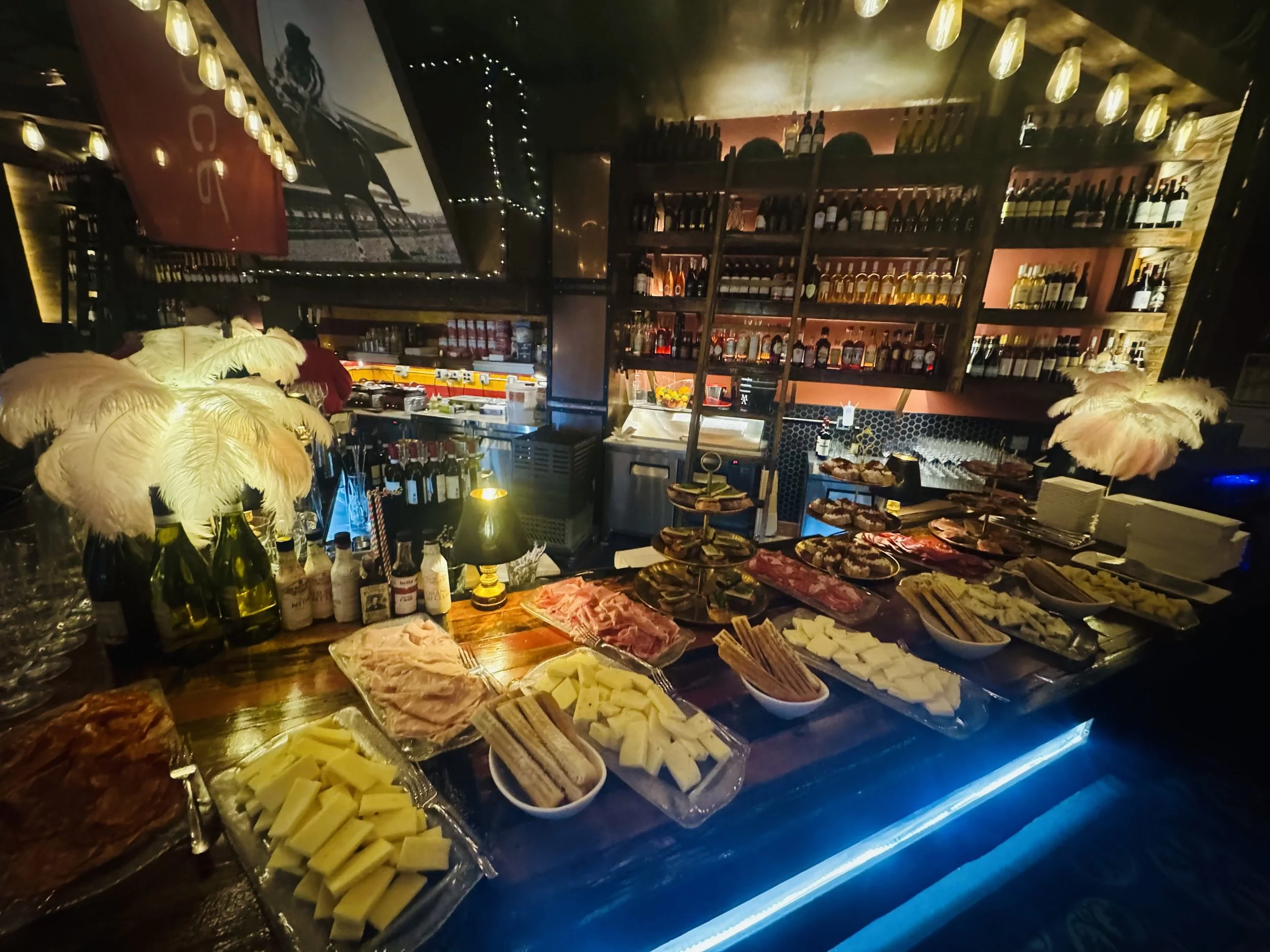 Wide view of the bar counter loaded with an aperitivo spread and feather centerpieces