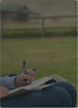 A hand writing in a journal outdoors in a fenced field