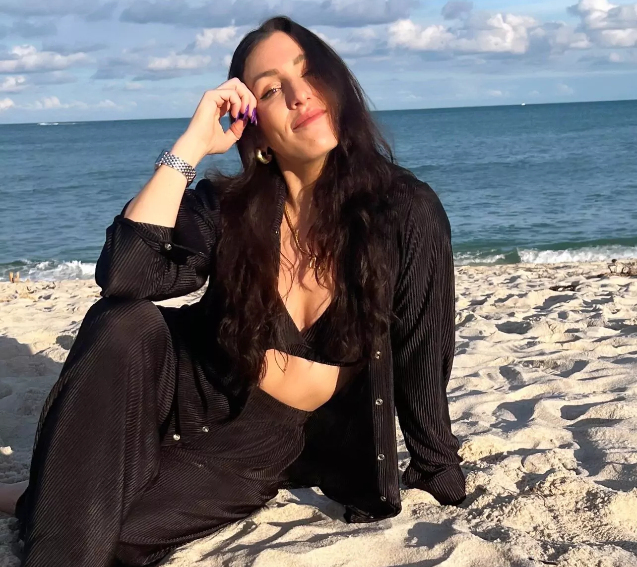 Lana Rose in black, sitting on a sandy beach with the ocean behind her