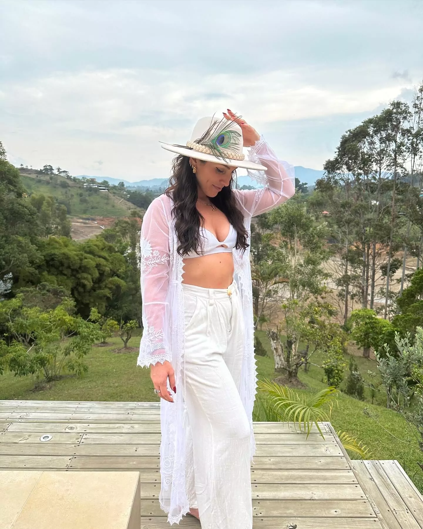 Lana Rose in white lace and a peacock-feathered hat standing on a wooden deck with a green hillside behind her