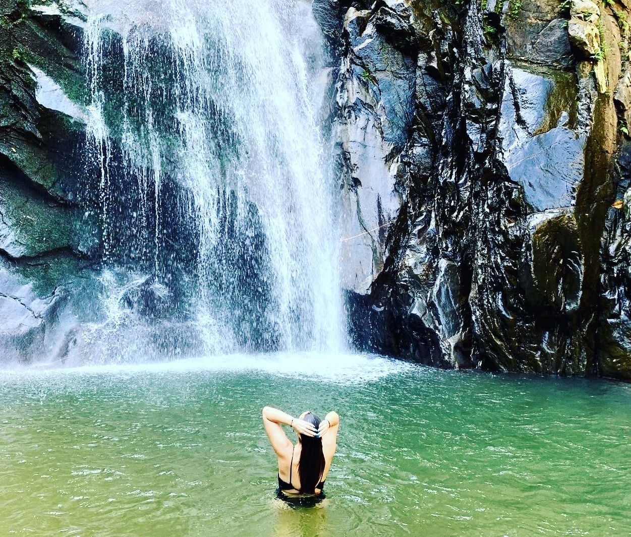 A woman standing waist-deep in a green natural pool beneath a large waterfall