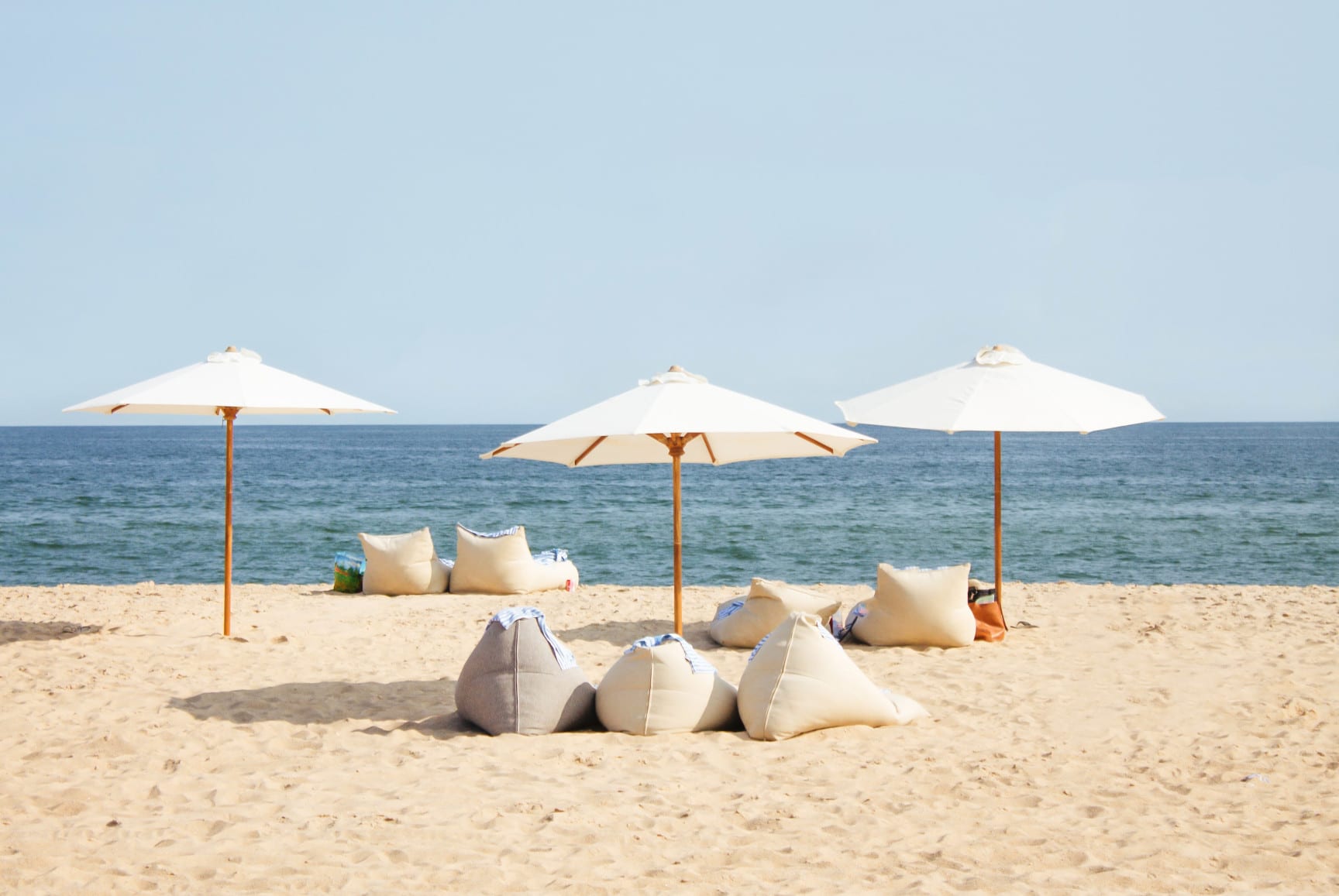 Three white beach umbrellas with linen bean-bag loungers on a sandy beach by the ocean